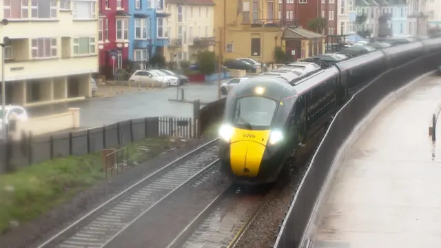 A train at Dawlish. The picture is slightly blurred as it appears to be raining. The lights are on. The train is moving towards the camera on the track. To the left of the tracks is a car park and buildings.