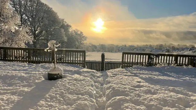 Thick snow surrounds a fence, the sun at a distance illuminates the area as snow can be seen in the landscape in the distance.