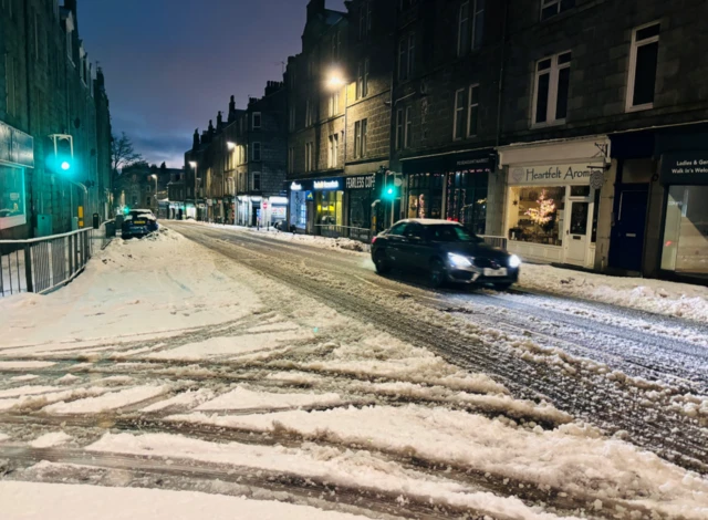 A car drives along a high street in snowy conditions. Parts of the road where the cars have been driven have been cleared