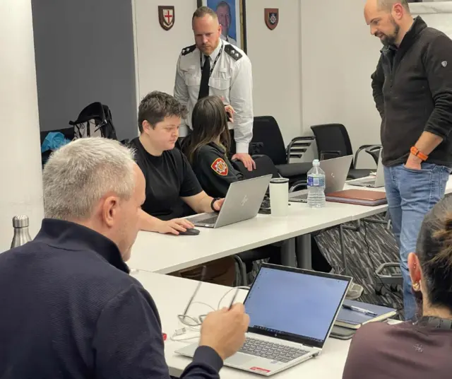 A picture of six people sat in a room. There are desks and computers with people stood around.