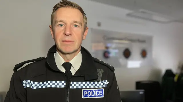 Jersey Chief Inspector in police uniform. He is looking directly at the camera and is stood in a control room. There are computers around and people sat at desks. The wall behind him is white and the background is slightly blurred.