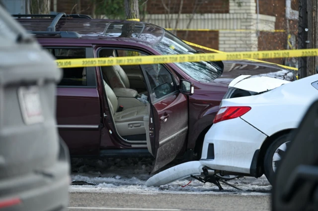 A dark burgundy SUV with an open passenger side door is cordoned off behind yellow police tape. Traces of blood can be see on the tan leather seats of the car