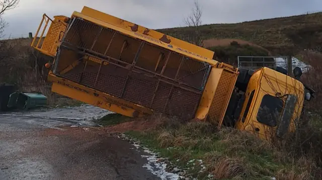 A yellow gritting lorry on its side with icy patches around it