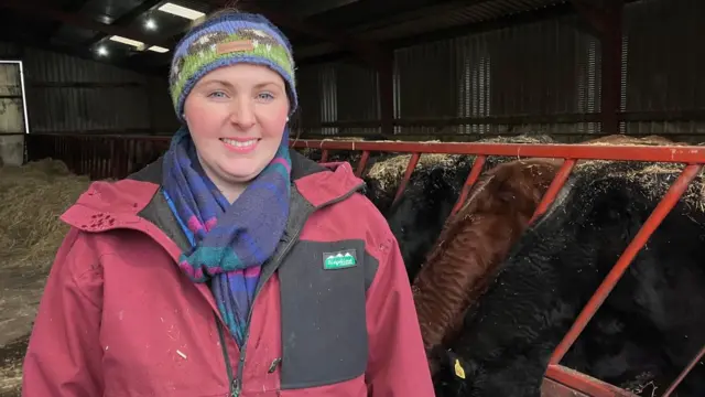 Farmer Katie Davies wears a red waterproof coat, a multicoloured scarf and hat as she stands beside some cows