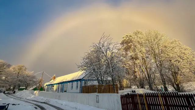 A railway is covered in snow next to a white and brown picket fence. A white 'fogbow' can be seen above a snow covered building and trees.
