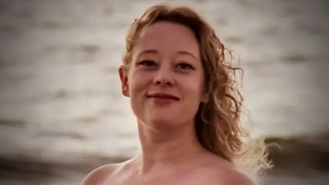A photo of Renee Nicole Good. She is standing on a beach with her back against the grey sea. Her hair is wavy blonde and she is smiling at the camera
