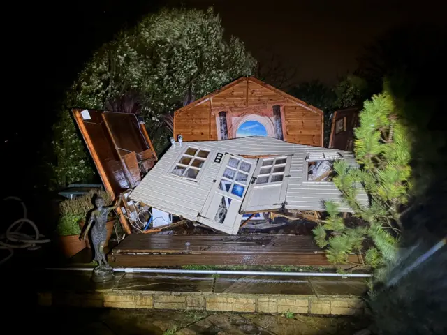 A picture of a shed damaged by the storm. It is wooden and white in colour.