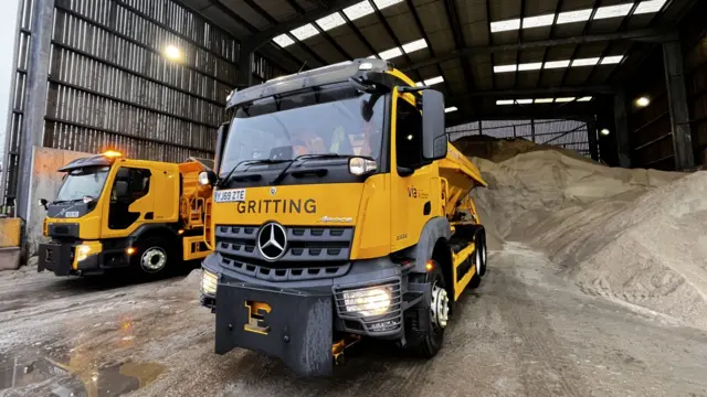 Two large yellow vans for gritting in a depot.