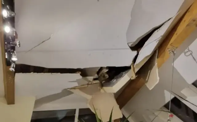 A picture of the roof. It is severely damaged and the image is taken from inside of the property. A set of lights can be seen to the left of the image