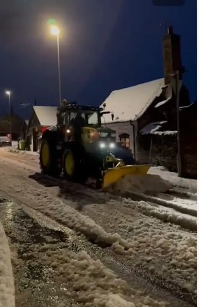 A tractor with a snow plough on the front drives down a snowy road
