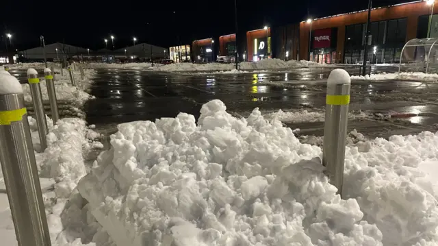 A pile of snow around bollards in front of a cleared retail park car park