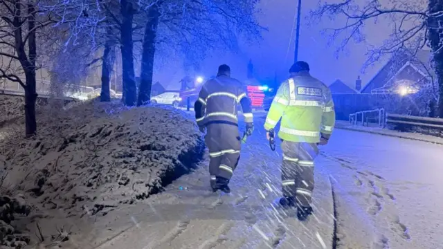 A rear view of two fire fighters in hi-vis uniform walking up a suburban street in heavy snow.