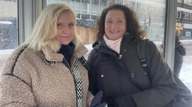 A woman with blonde hair and light brown fluffy coat stands next to a woman with brown hair and a black coat. They're both smiling while they stand under a bus stop shelter. A snow-covered road is visible behind them.