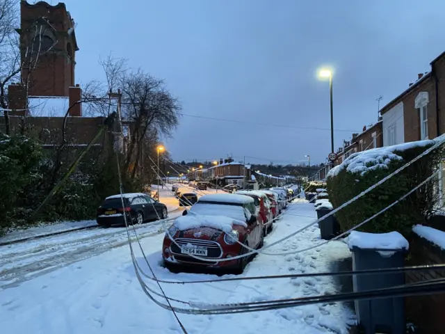 A telegraph pole has partially fallen over at the side of a snow-covered residential street. The wires are hanging down across a red mini cooper parked at the side of the road.