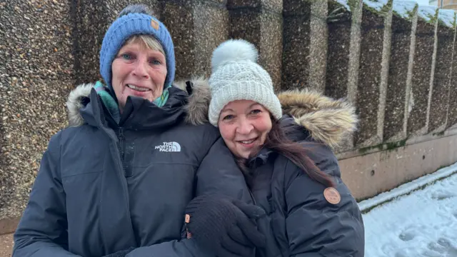 Two women wearing bobble hats and large black coats, smile as they stand on a pavement with their arms linked.