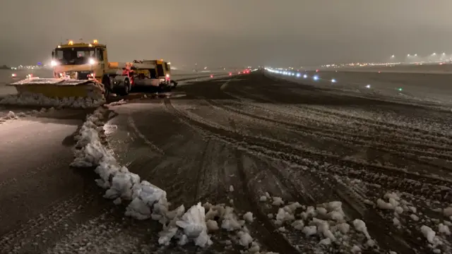 Vehicles parked up at the side of an airport runway, which is covered in snow. A vehicle can be seen moving snow away from the runway.