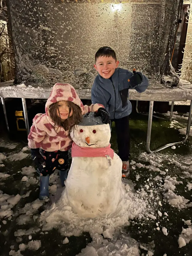 A boy and a girl pose for a picture while standing behind a snowman they have just built.