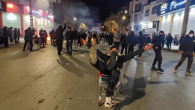 A protester in black cap and with his face partially covered up sits in a shopping cart with his arms and right leg extended. Behind him, several men stand together scattered across a commercial street, also with their faces covered