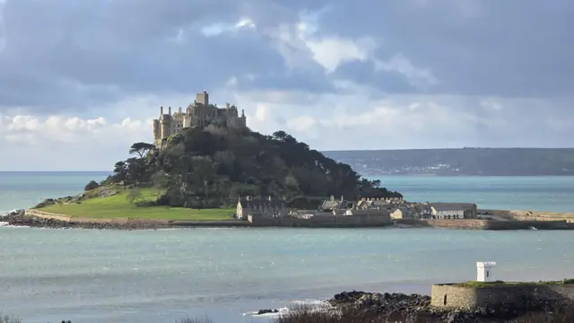 A picture of St Michael's Mount. It is a large building on top of a mountain surrounded by a large number of trees.