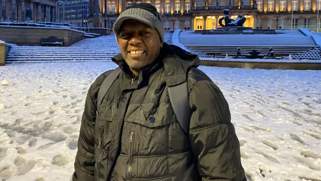 A man wearing a hat, a black coat and a backpack, smiles as he stands in a snow-covered city centre.