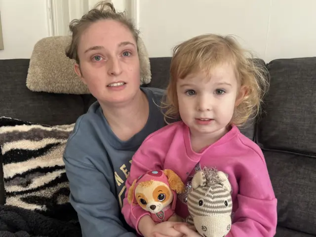 A picture of Emily and her daughter. She is wearing a blue jumper and her daughter is wearing a pink jumper and holding two toys.