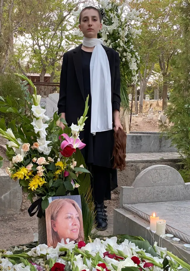 Roya Piraei poses next to her mother's grave. She has a shaved head and wears a white shirt and black trousers. in her hand she grips what looks to be the hair she shaved off. white lilies cover the grave, and a photo of Minoo, her mother, can be seen.