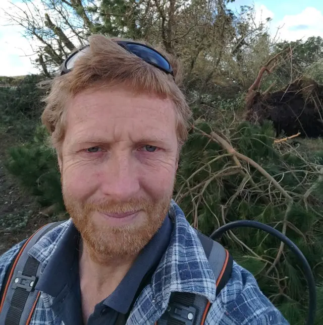Man stands in front of felled trees with sunglasses on his head
