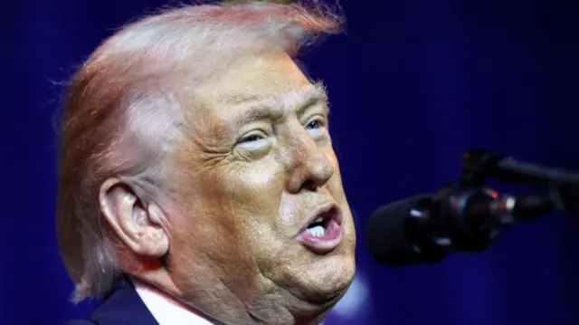 Close up of US President Donald Trump. He's speaking into a black mic, photo cuts off just below the neck, partially showing a white shirt and blue suit jacket
