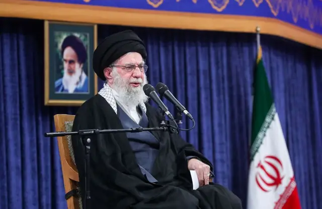 Ali Khamenei in black vest and grey robes sitting down on a wooden chair. Behind him, a frame with a picture of Supreme Leader Khomeini and an Iranian flag to his left