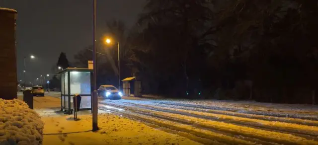 A snow covered road with a car with headlights on driving by