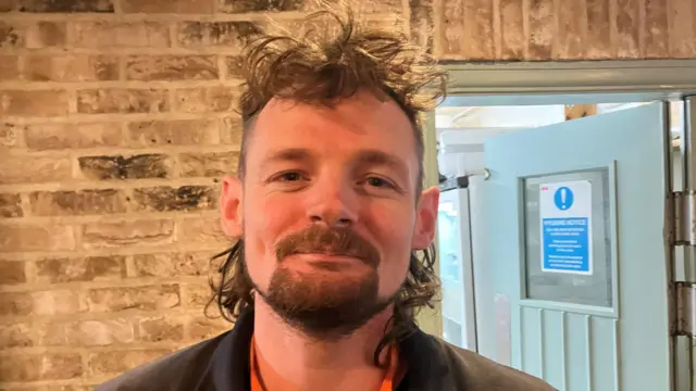 A picture of Kai Johnson, the supervisor at the Heron Inn. He has long hair and a brown beard.