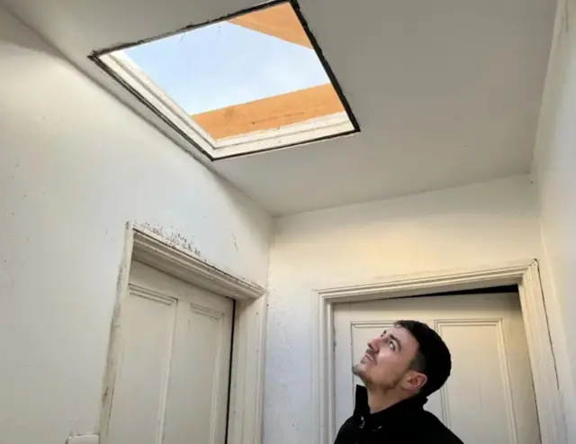 A man is pictured looking up towards a large hole in his roof. He has black hair and is stood in his hallway.