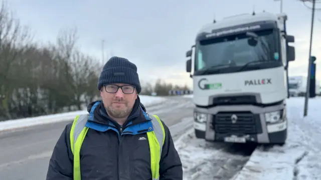 Man wearing glasses and hat stood next to HGV stuck in snow