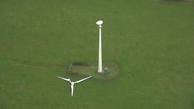 A wind turbine with its blades which are on the grass away from the main turbine.