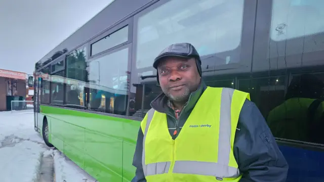 A man wearing leather flat cap and high vis vest stood next to a bus