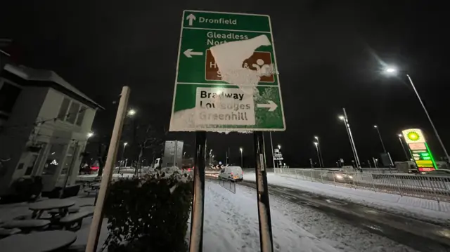 A roadsign partially obscured with snow in front of a road which is partially covered by traffic appears to be moving.