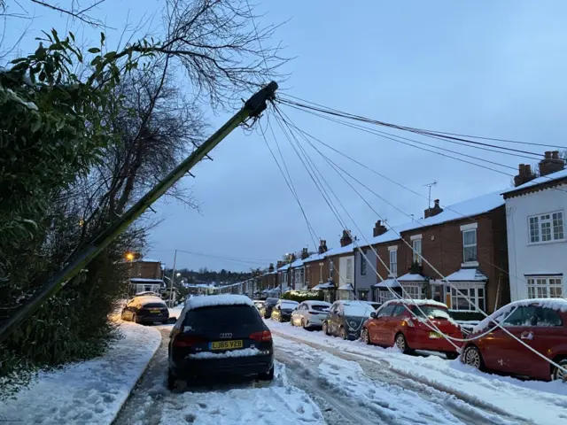 A telegraph pole at the side of a snow-covered residential street has partially fallen and is learning at an angle. Its wires are sagging down towards the street.
