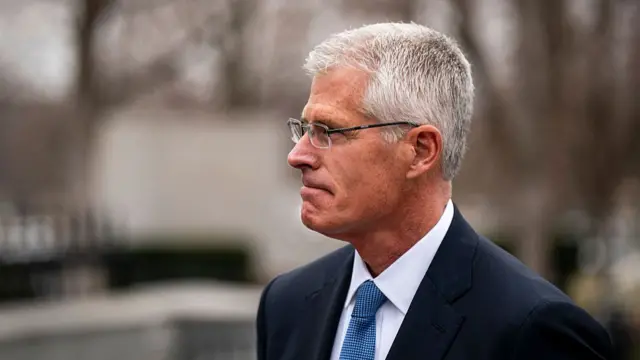 A man with gray hair and glassess wearing a navy suit and lue tie looks serious off to the left