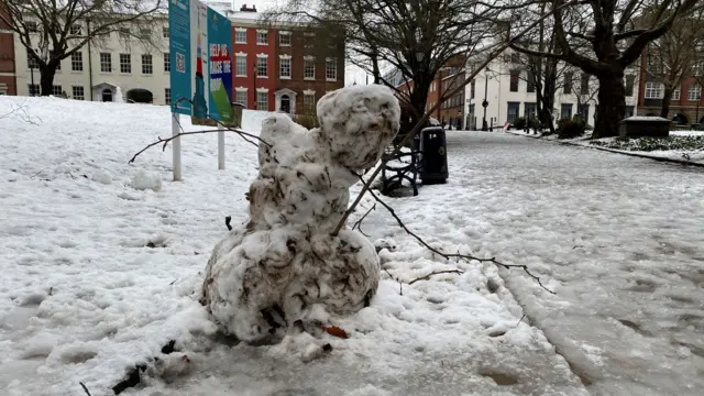 A snowman leaning to one side, with branches poking out of it, in the middle of a park, which is covered in snow and ice.