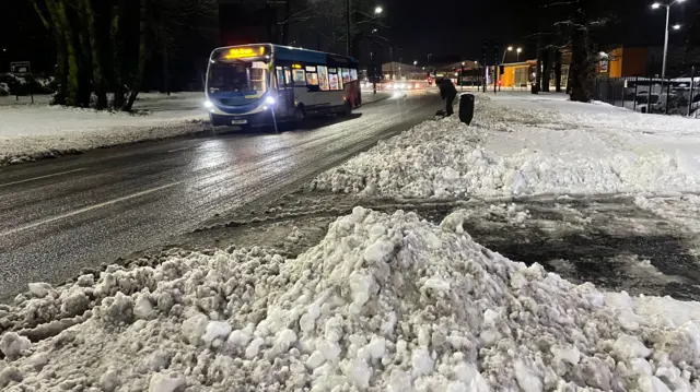 A pile of snow sits near to a cleared road with a bus travelling along it