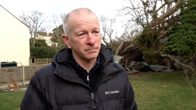 James Robinson looking to the right and speaking with a reporter. Behind him are fallen trees and he's wearing a black coat.