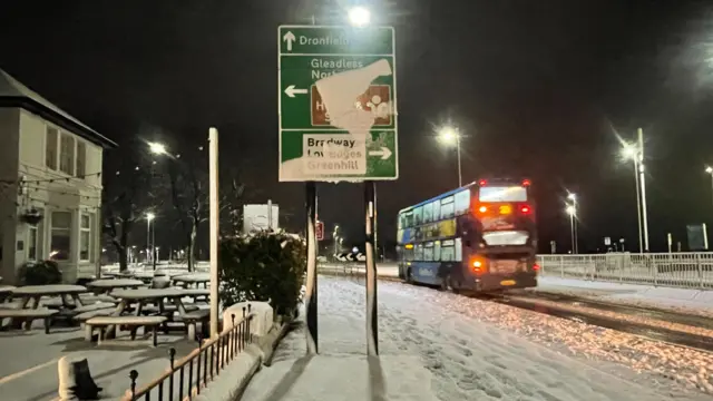 A roadsign partially obscured with snow in front of a road which is partially covered by traffic appears to be moving.