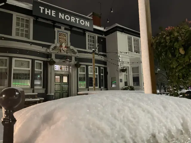 Snow covered car roof in front of a pub where there is a couple of inches of snow in a beer garden