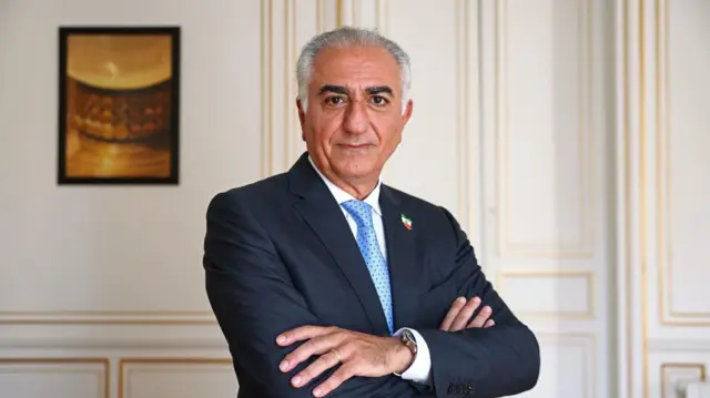 A man with dark eyebrows and white hair wears a dark suit and fold his arms while looking at the camera.