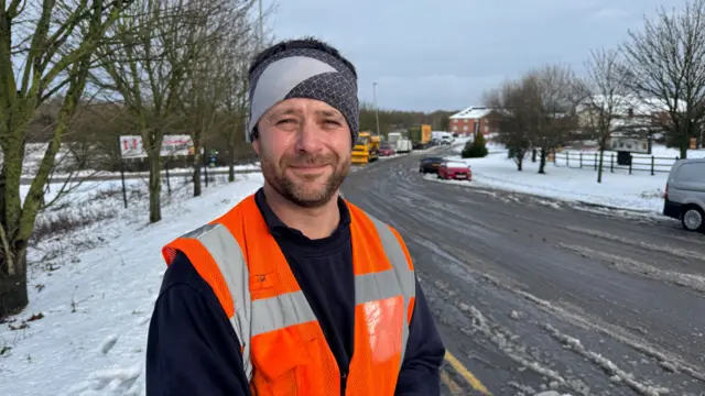 A man in a orange hi-vis jacket stands in front of a snow covered road
