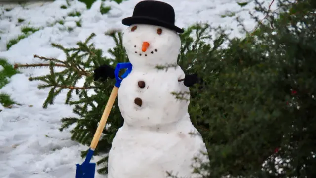 A snowman wearing a black bowler hat and has a blue spade leaning up against it.