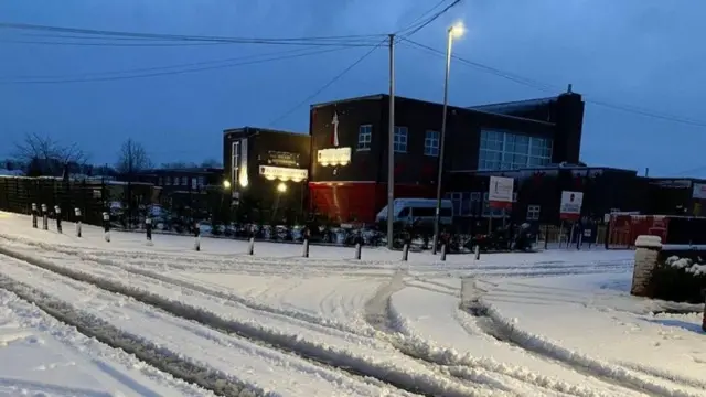 A snow-covered road leading to the entrance of a school. The large school building can be seen behind a fence and next to a car park.