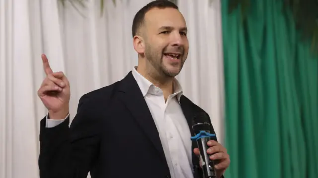 Zack Polanski speaking into a mic wearing a black suit