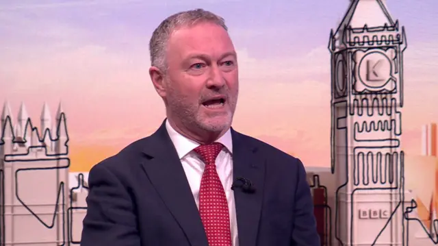 Steve reed in black suit, red tie and white shirt sitting down inside a TV studio