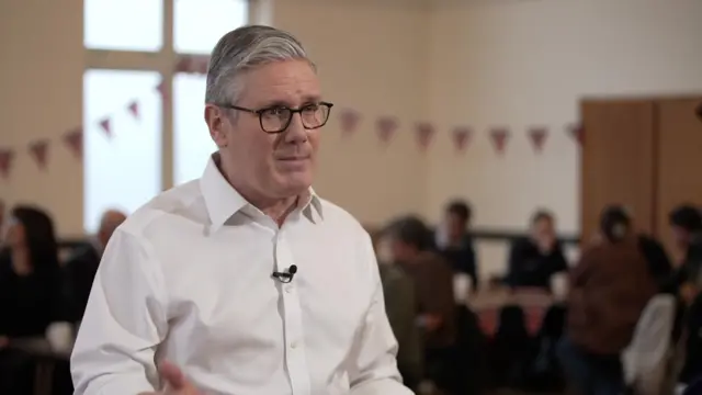 Keir Starmer mid-speech in white shirt in a room with other people sitting down behind him